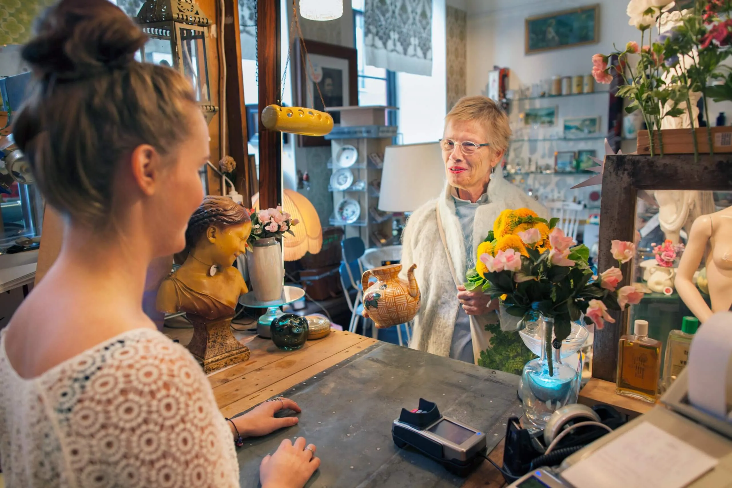 A happy elderly woman stands at a flower shop counter, smiling beside a vase filled with vibrant flowers