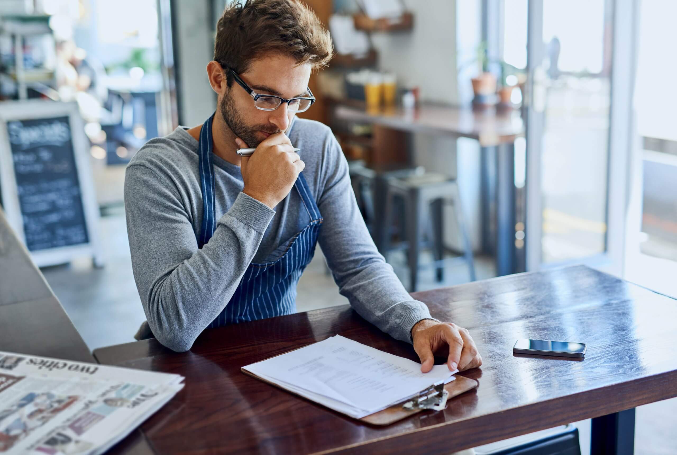 A man wearing glasses is seated at a table, writing with a pen on a piece of paper