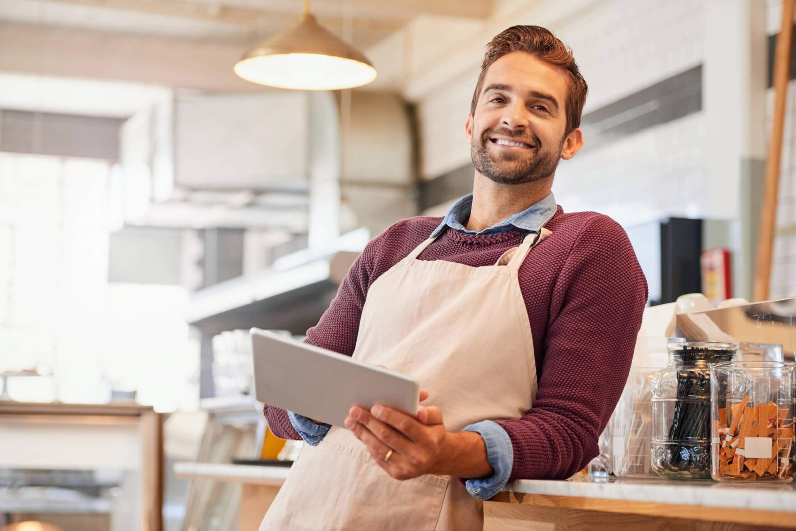 A cheerful man in an apron stands holding a tablet, exuding warmth and hospitality in a culinary environment