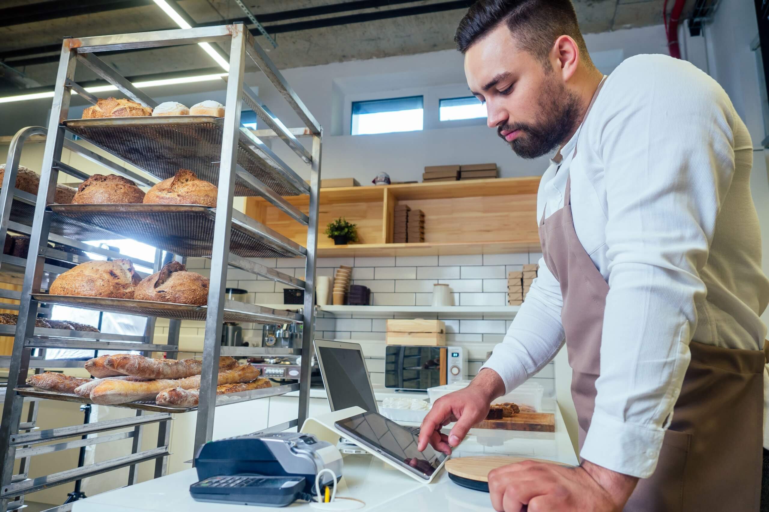 Male baker in white shirt and apron using laptop computer for POS and kitchen display screens management next to bread display rack in modern bakery kitchen