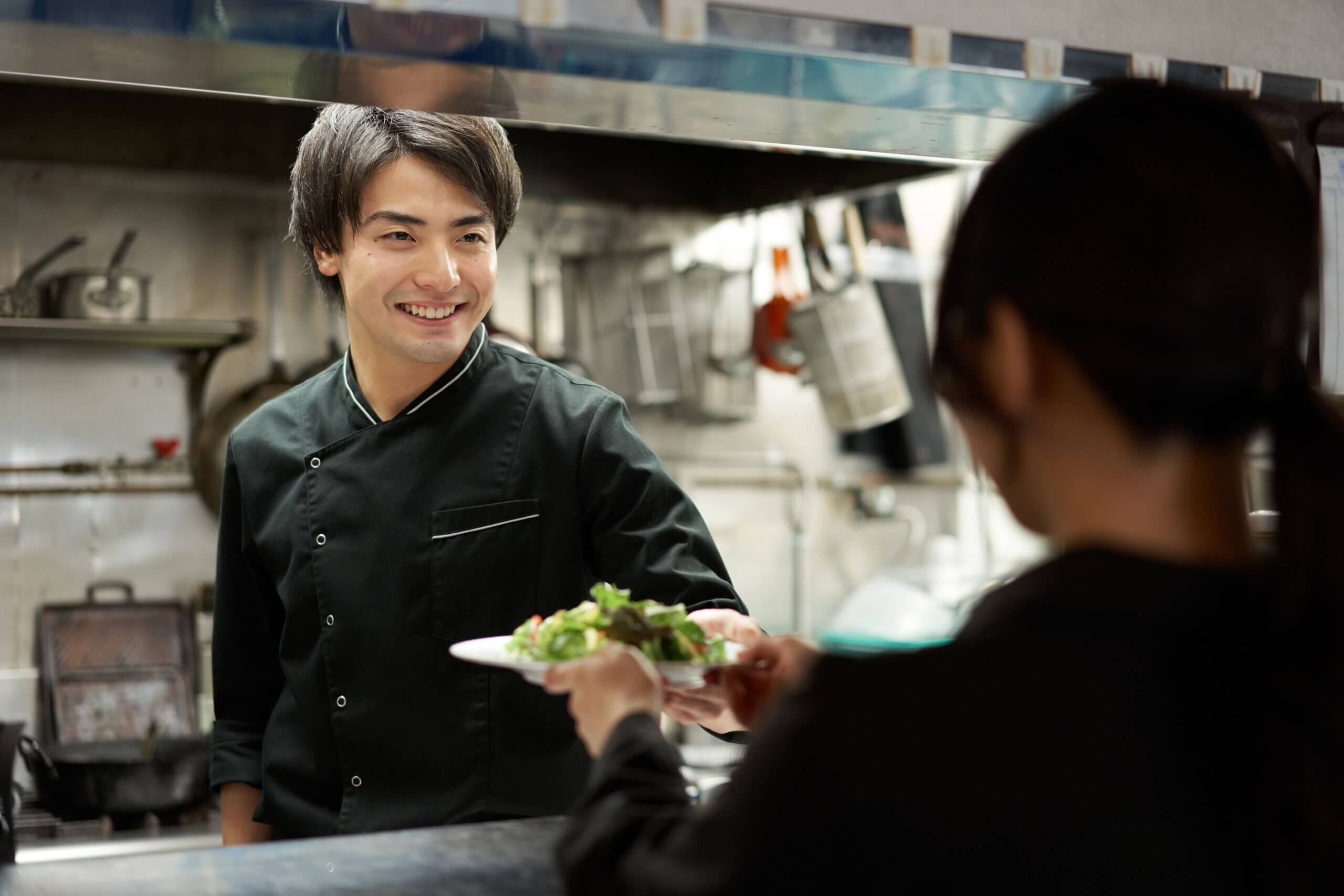 A happy chef handing a prepared meal to a server in a restaurant kitchen, highlighting seamless service with POS & kitchen display screens.