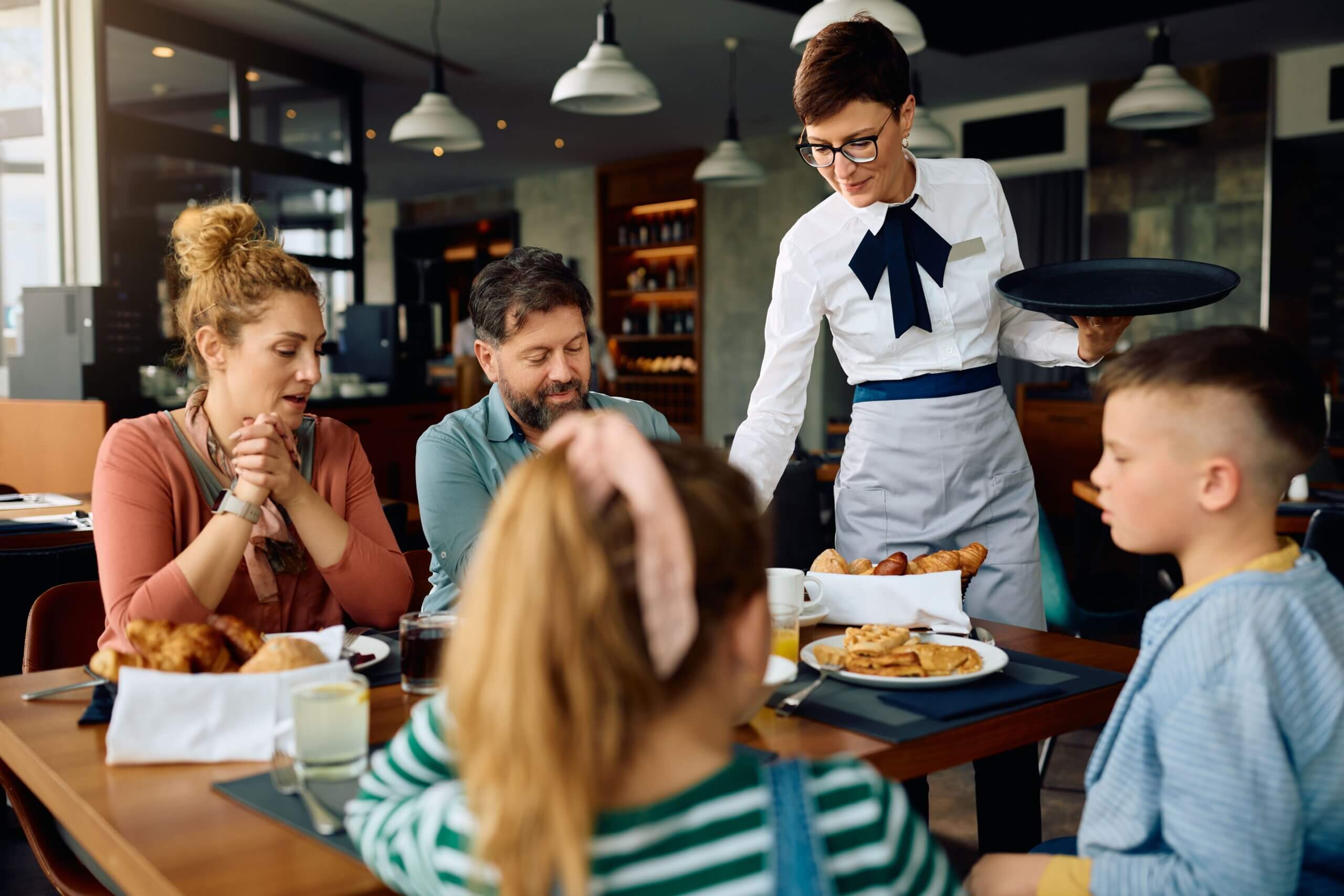  smiling waitress serving breakfast to a family at a restaurant, with fresh pastries and pancakes on the table.