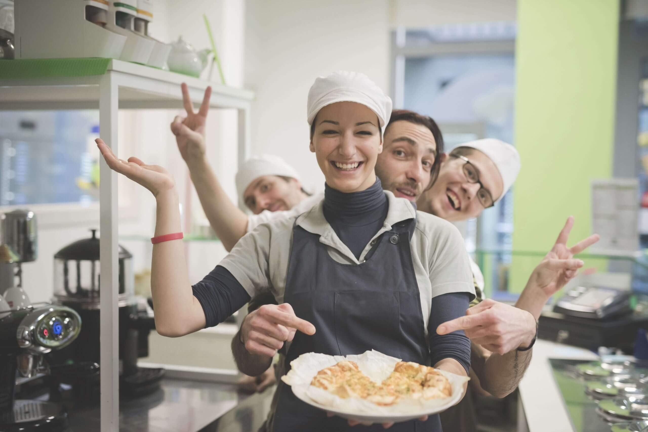 Cheerful kitchen staff in white chef hats and aprons celebrating and making peace signs while holding a plated dish in a bright commercial kitchen