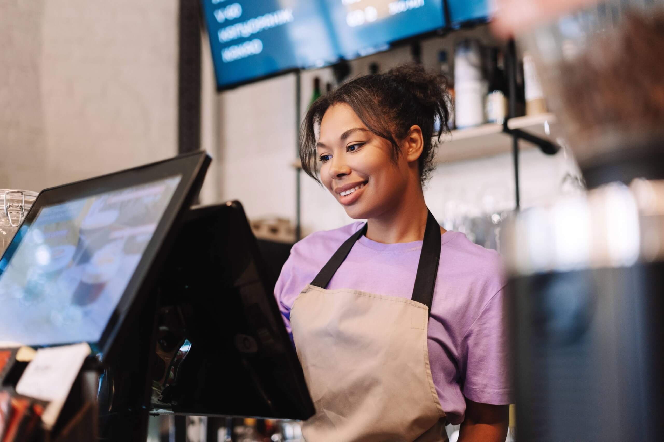 Smiling female cashier in purple shirt and apron using POS & kitchen display screens system at modern restaurant counter with digital menu boards visible in background