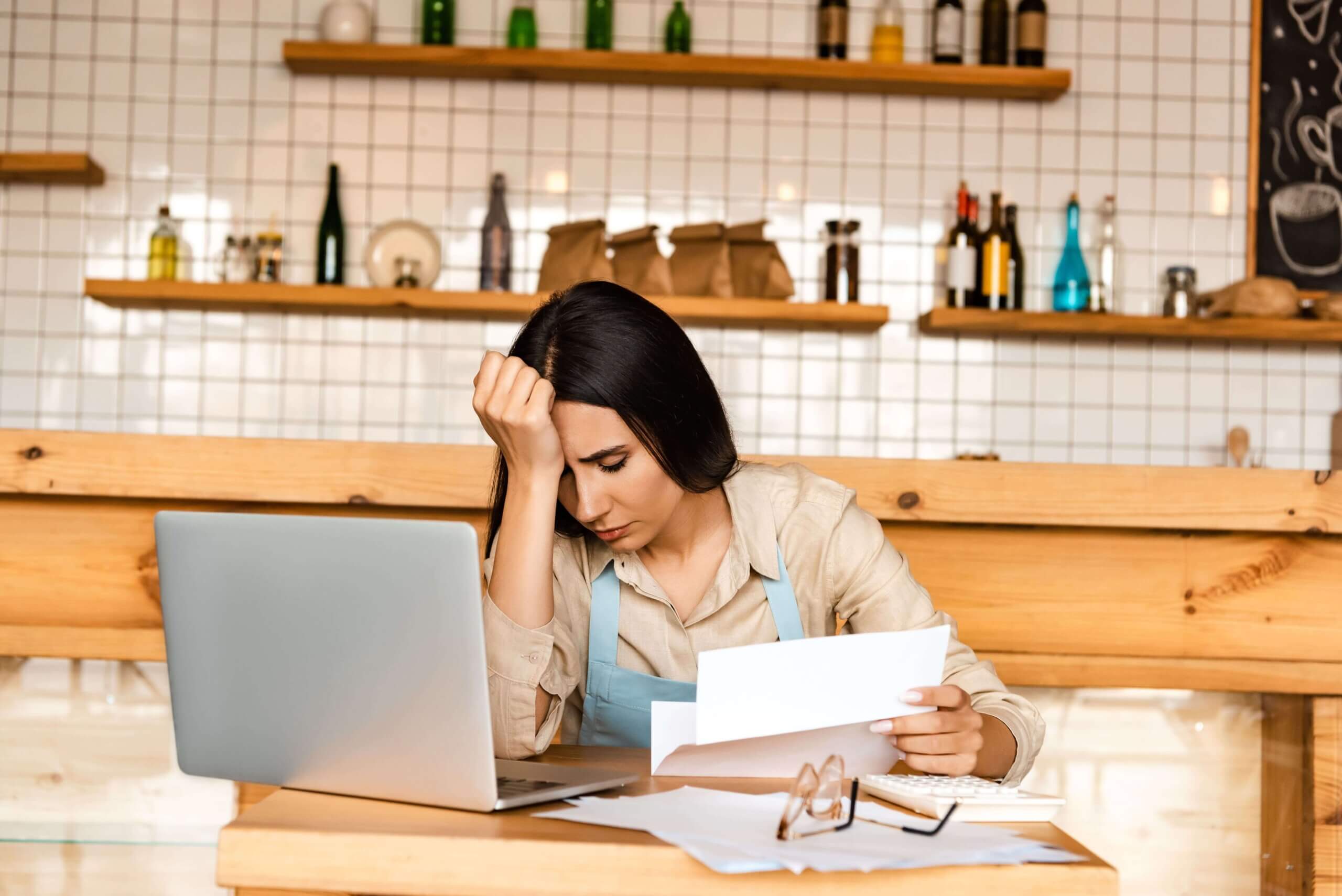 Female restaurant owner in apron sitting at wooden counter reviewing financial reports and using laptop, likely analyzing restaurant POS system data for business management