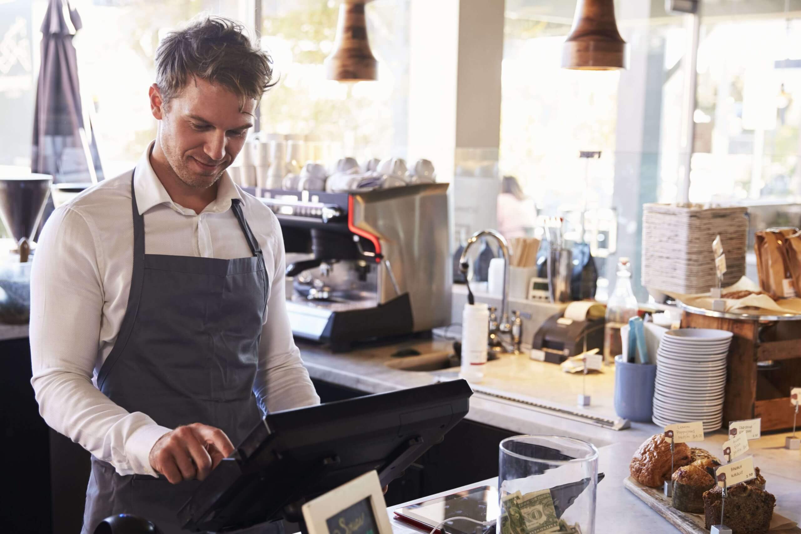  Male server in apron using restaurant POS system touchscreen terminal to process customer orders at modern cafe counter with pastries and coffee equipment
