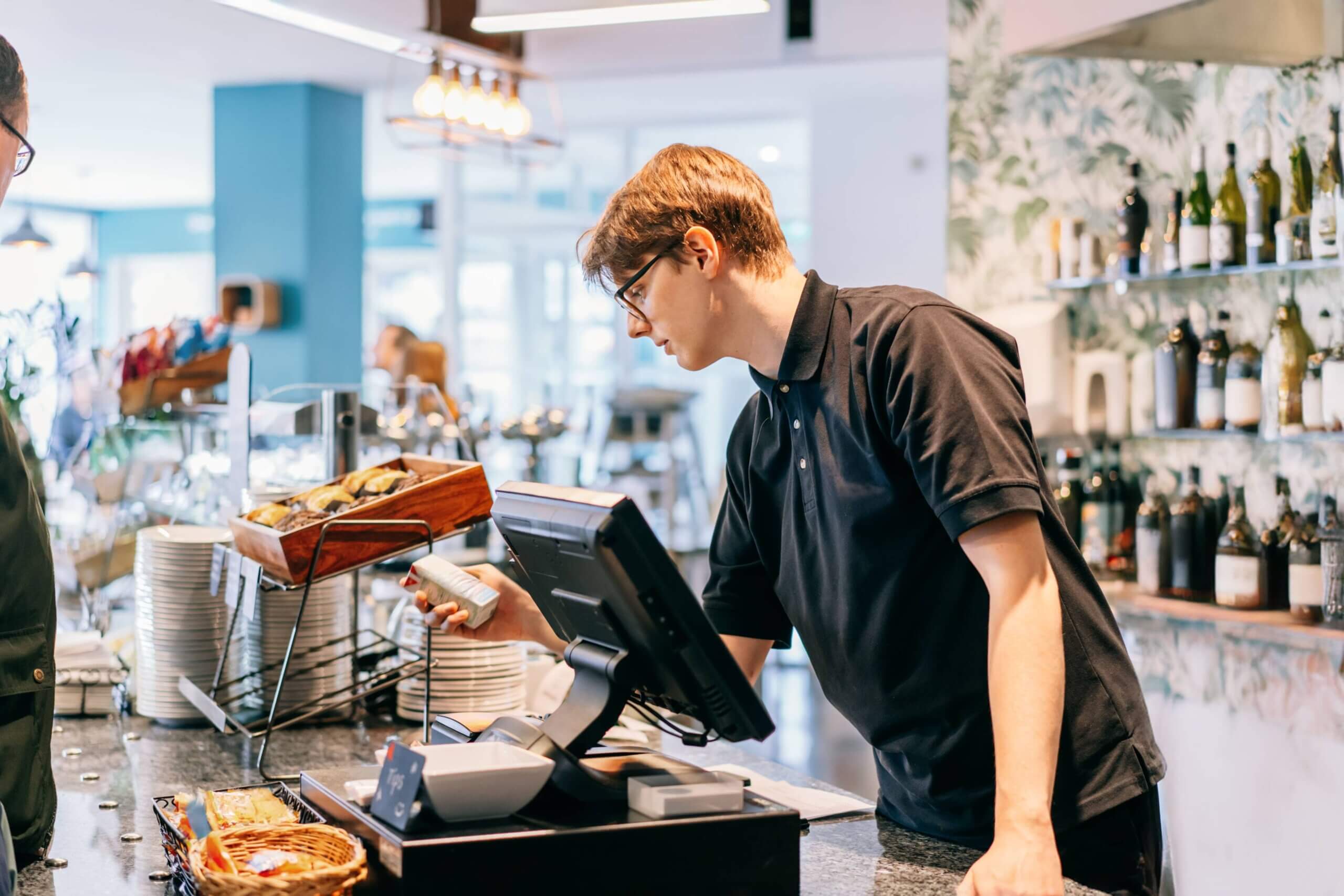 Male server in black polo shirt scanning product barcode with handheld scanner at restaurant counter with plates and bar setup in background
